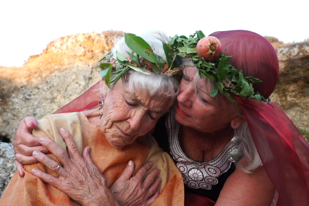 Two elderly women wearing leaf and fruit crowns, embracing outdoors, with one woman comforting the other.