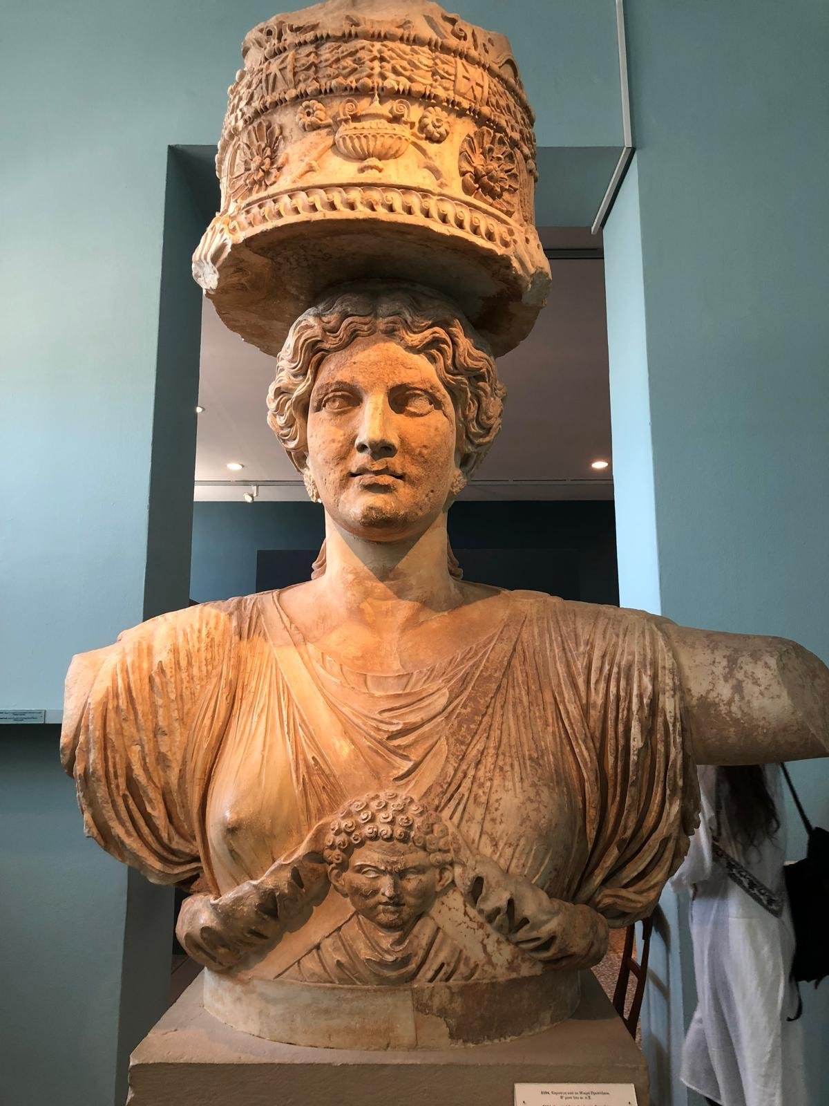 Ancient statue Demeter wearing an intricate headpiece and draped clothing, displayed in a museum setting. The statue has detailed carvings and a decorative element on the chest.