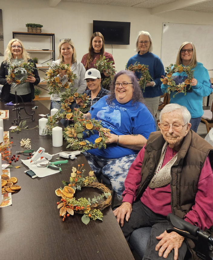 Group of women and an elderly man making fall wreaths with dried oranges and flowers in a crafting room.