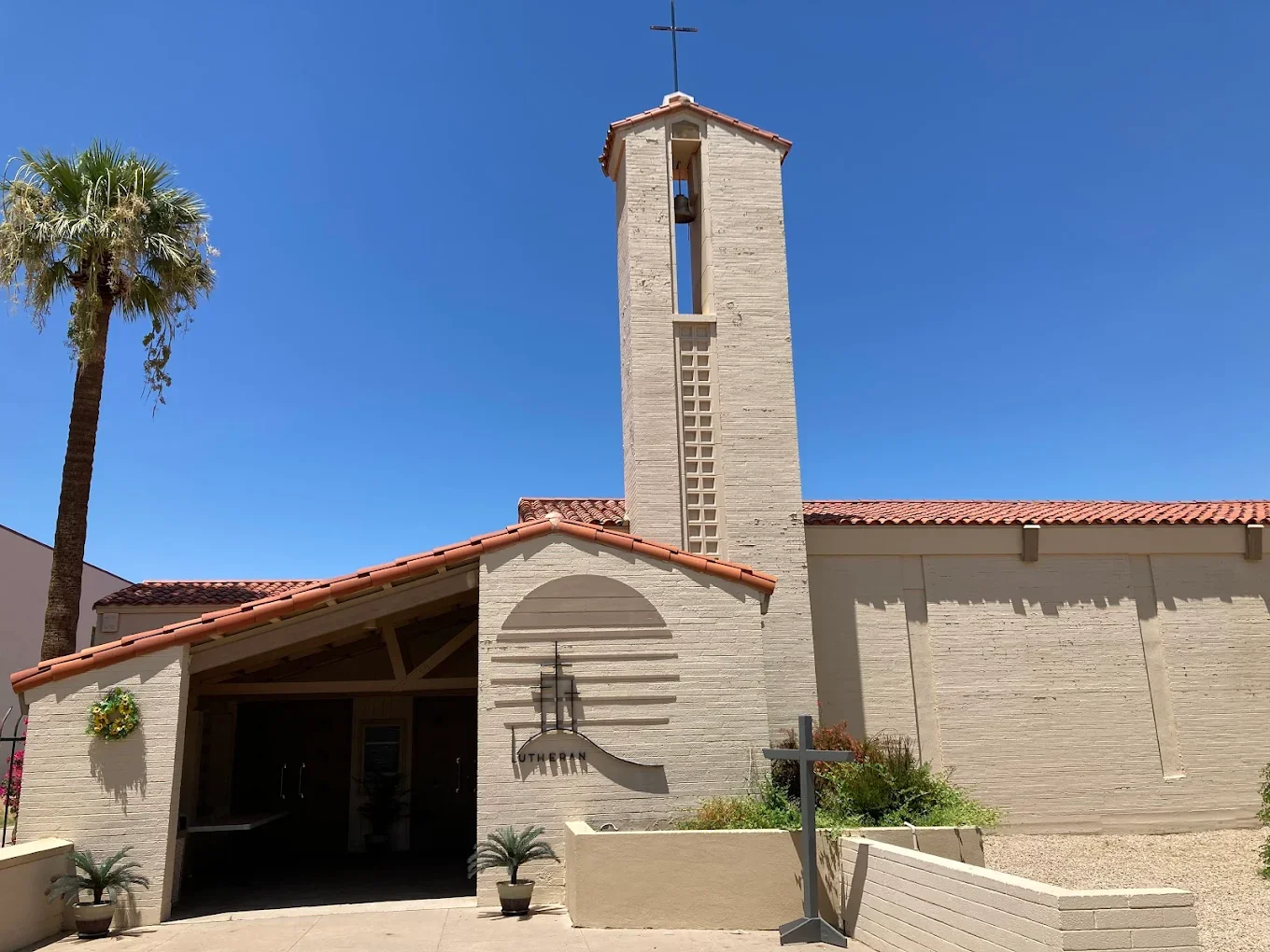 A church with a tall bell tower, a palm tree to the left, desert plants, and a sign that reads "Lutheran" at the entrance, under a clear blue sky.