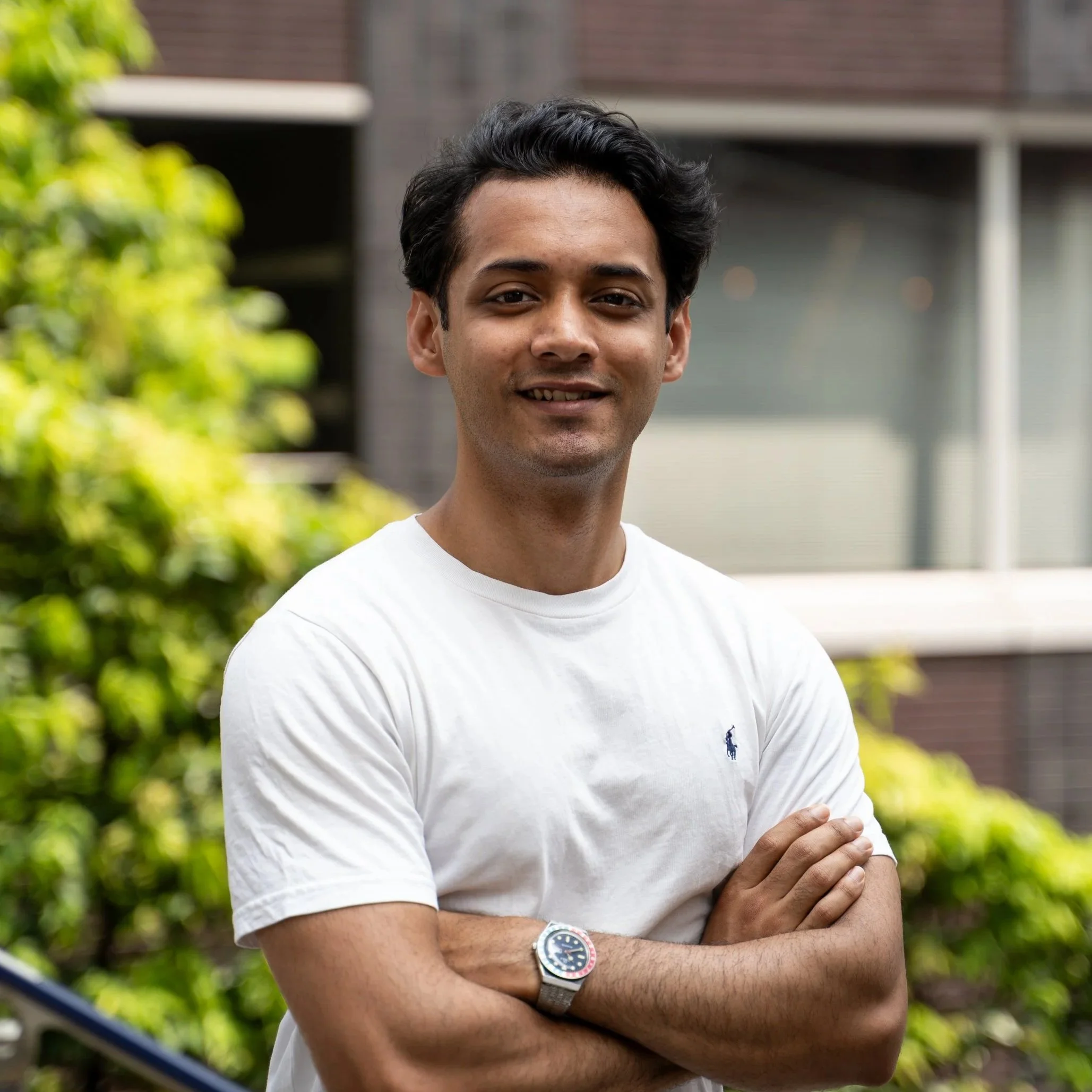 A young man with dark hair smiling, wearing a white T-shirt and a wristwatch, standing outdoors near some green foliage and a building in the background.
