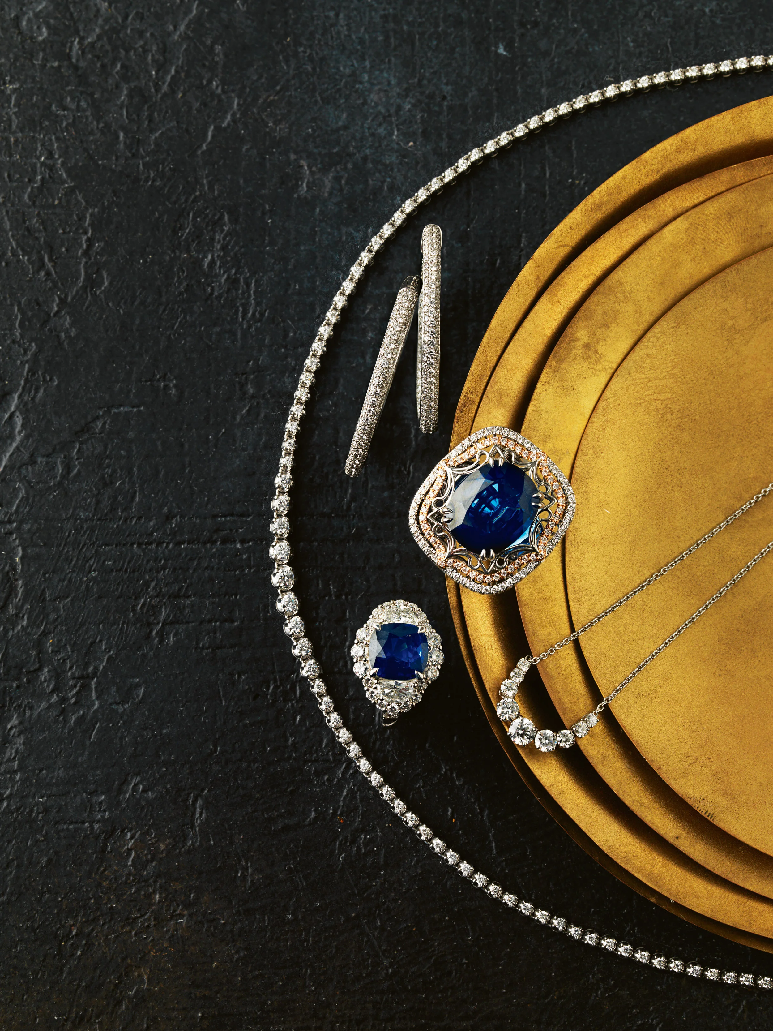 Jewelry set with blue gemstones, including a necklace, earrings, a ring, and a bracelet, displayed on a black textured surface and a golden tray.