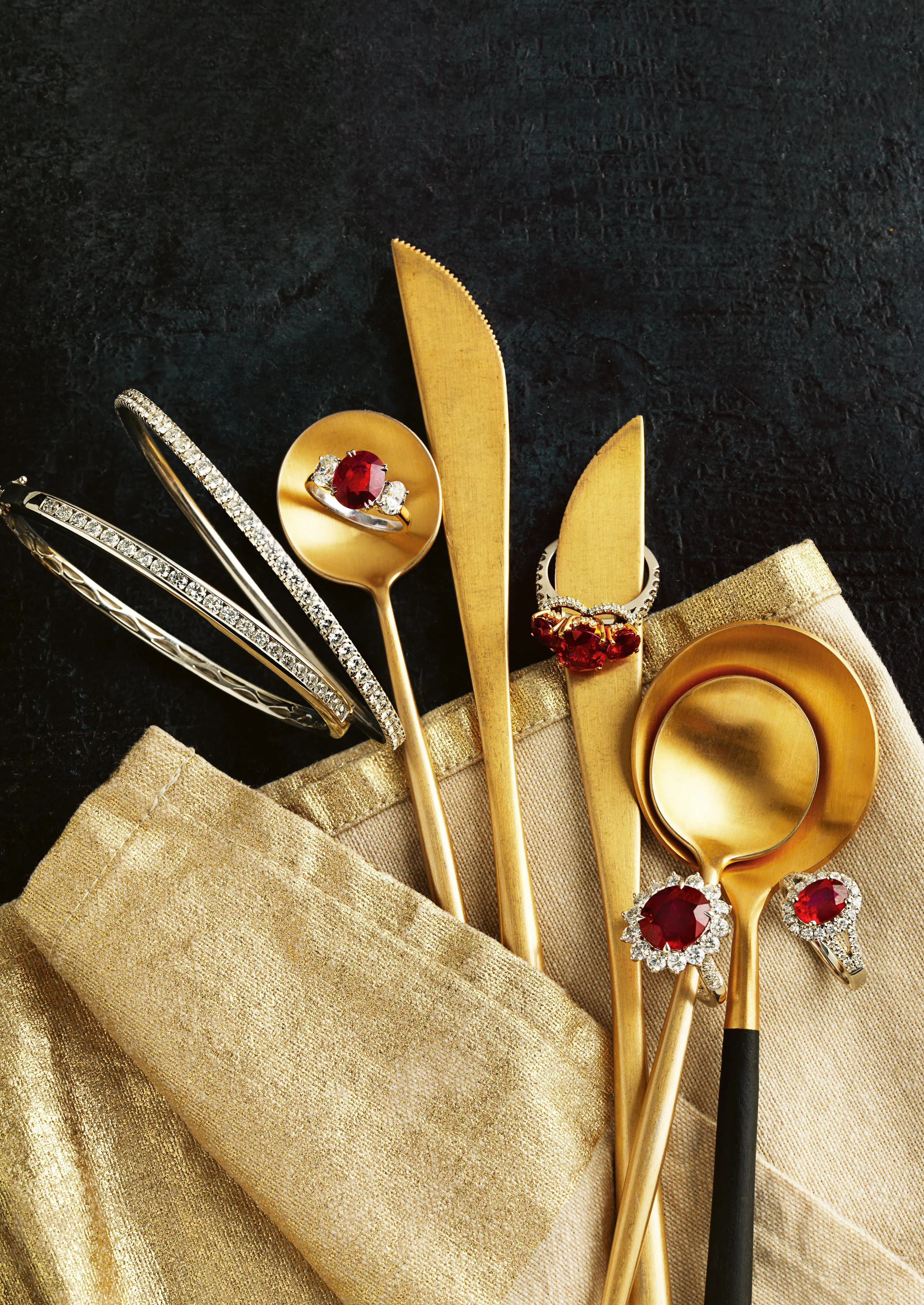 Gold-colored serving spoons and jewelry with red gemstones, set on a gold cloth and black surface.