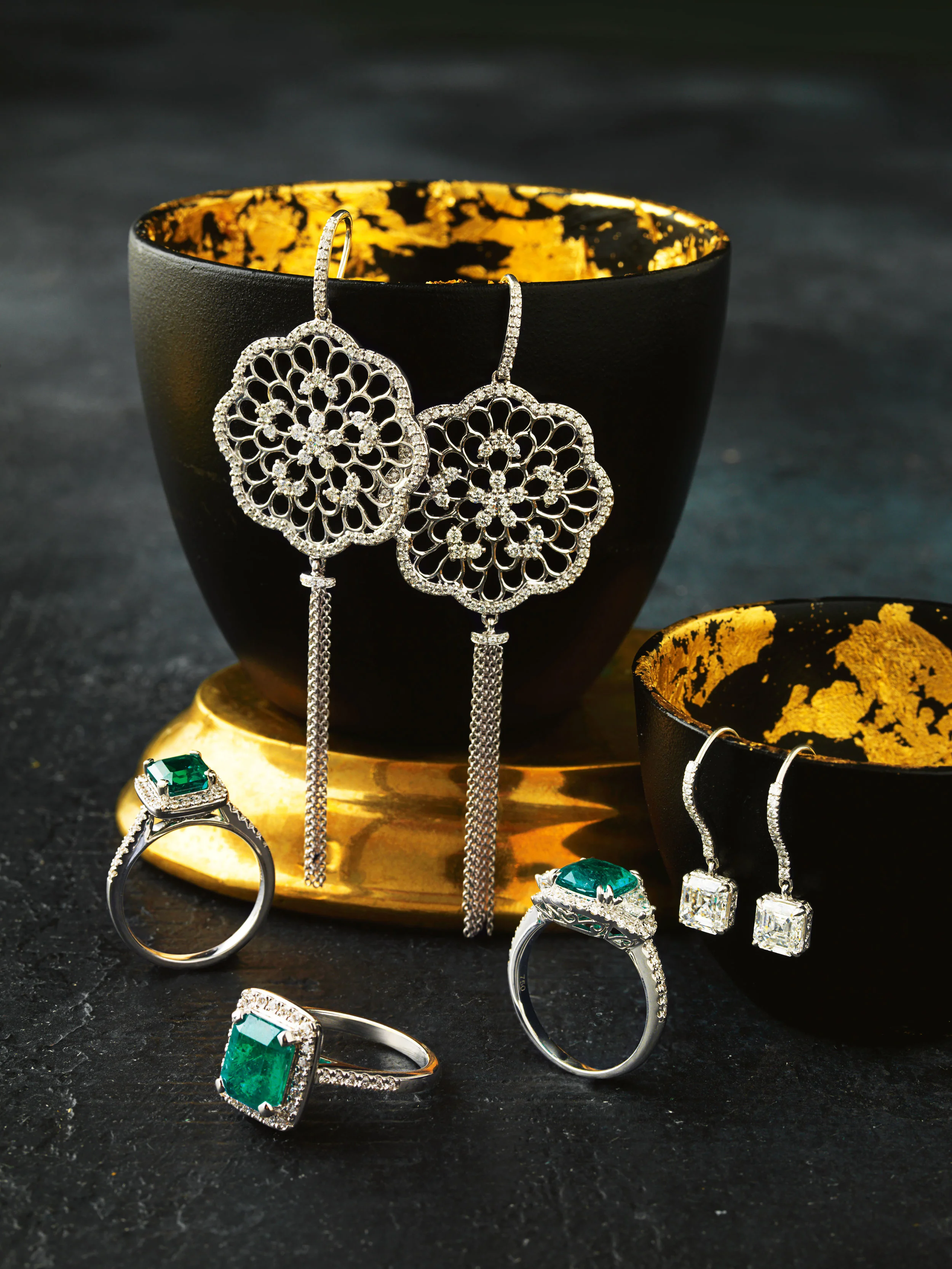 Elegant jewelry including two pairs of diamond earrings, two diamond rings, and a green gemstone ring displayed on black bowls with gold interior accents.