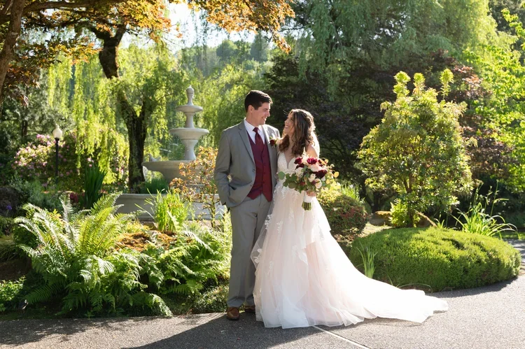 A bride and groom standing under a tree in a garden, smiling at each other. The bride is holding a bouquet of flowers, wearing a white gown, and the groom is dressed in a gray suit with a maroon vest and tie. There is a fountain behind them and lush 