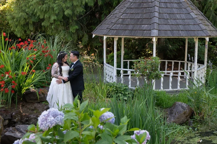 A bride and groom standing close and smiling in a lush garden with a white gazebo and colorful flowers.