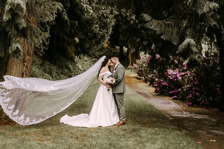 A bride and groom sharing a close moment in a lush garden, surrounded by large evergreen trees and vibrant pink flowers.