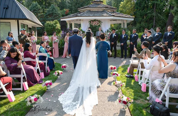 Outdoor wedding ceremony with bride and groom walking down the aisle as guests look on and take photos, surrounded by flowers and greenery.