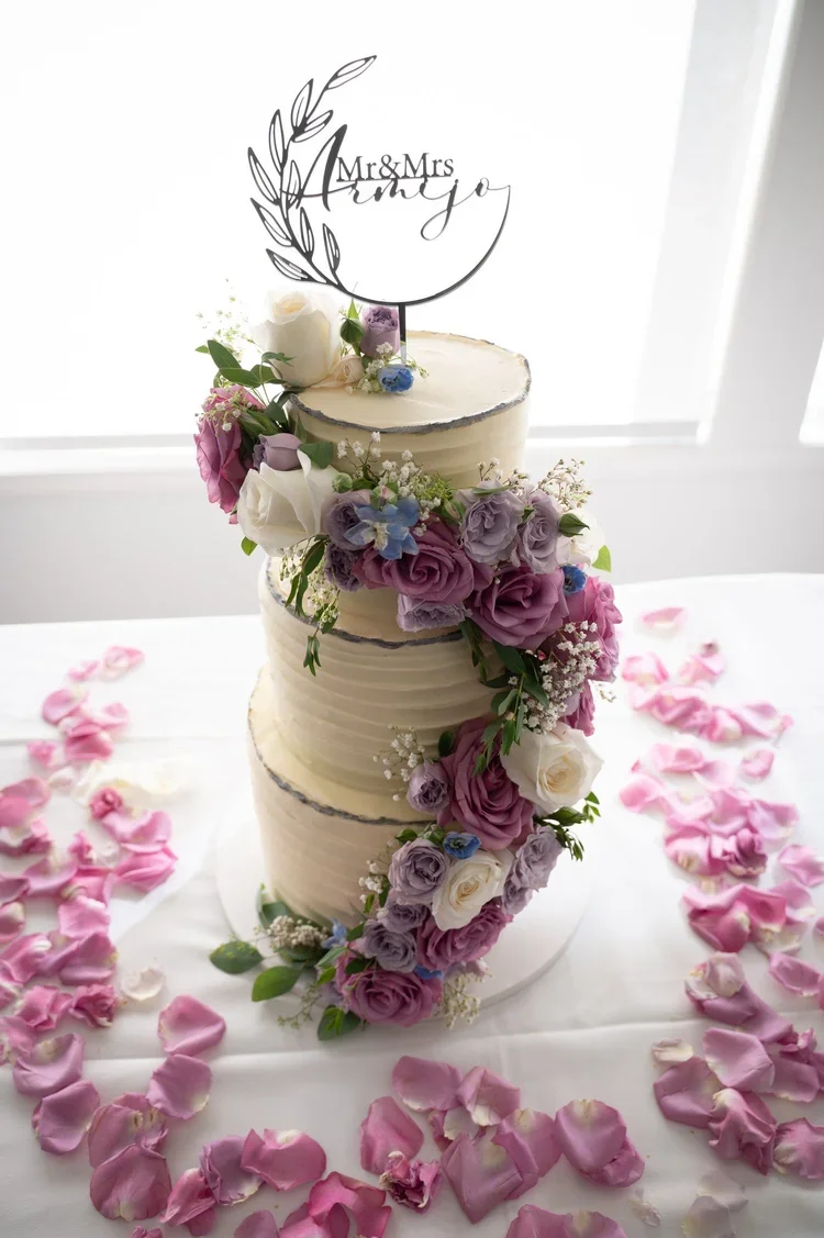 A multi-tier wedding cake decorated with fresh flowers including roses, lisianthus, and baby's breath, topped with a "Mr & Mrs" cake topper in a moon and leaves design, surrounded by scattered pink and white rose petals.