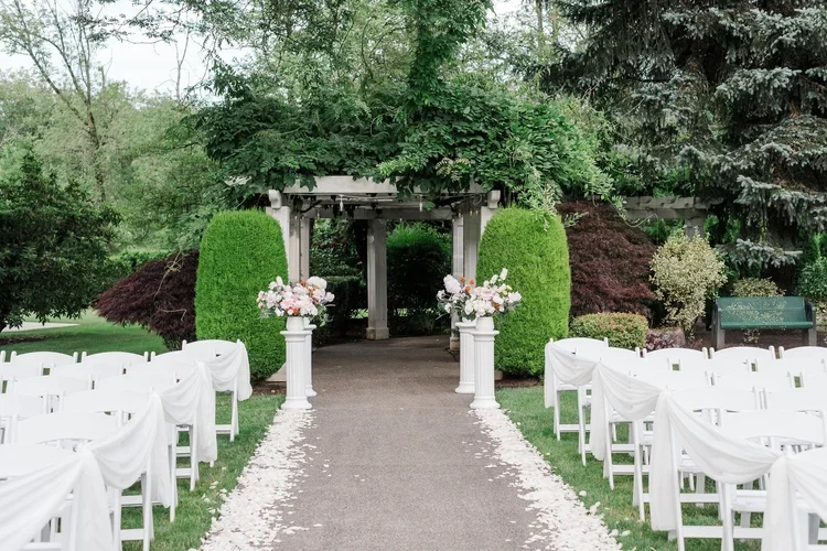 Outdoor wedding ceremony setup in a garden with white chairs, a floral aisle, a white gazebo adorned with greenery, and flower arrangements.