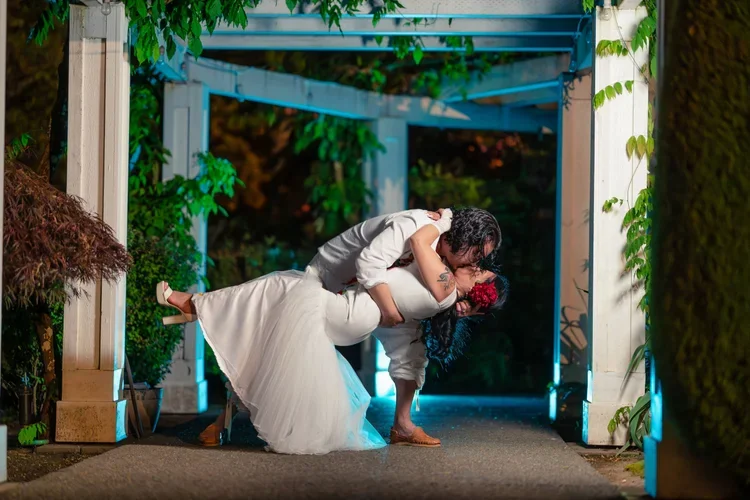 People sharing a romantic kiss at night under a decorated archway, with one person holding a bouquet of red roses, surrounded by greenery.