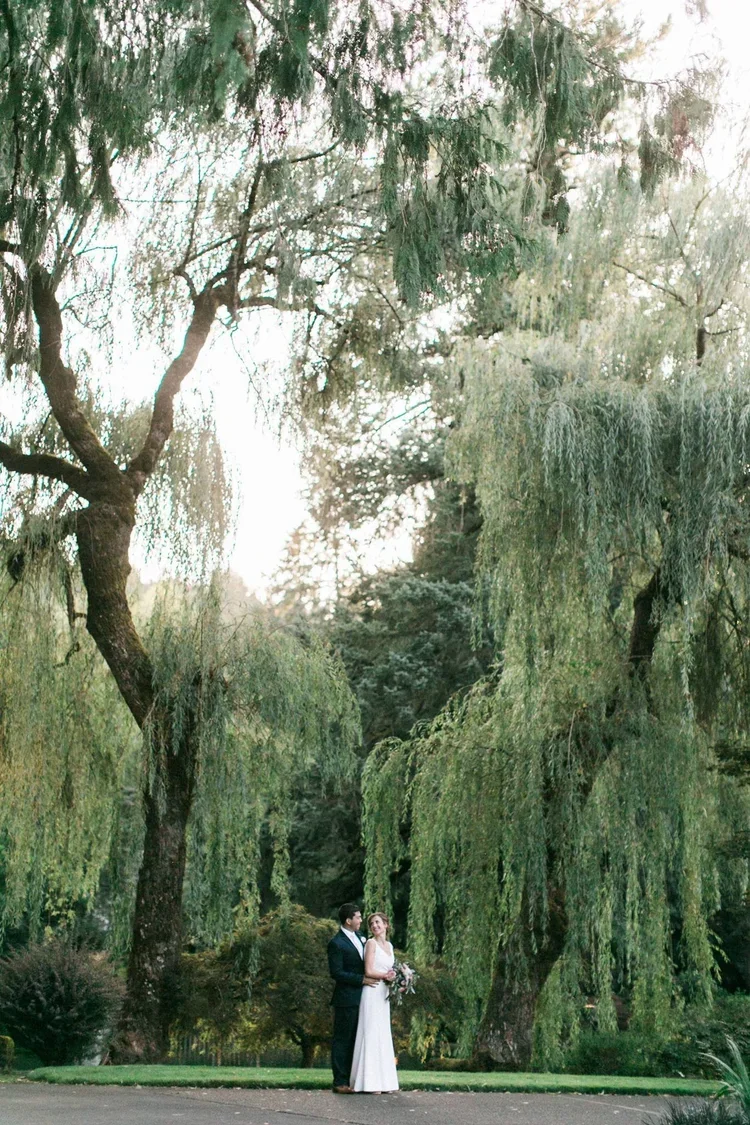 A man and woman in wedding attire standing in a lush green park beneath large, hanging willow trees.