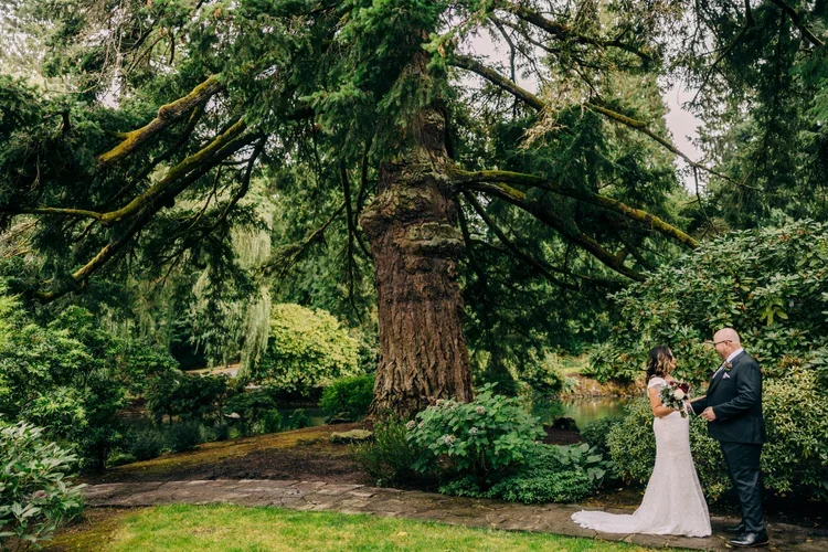 A bride and groom standing outdoors in front of a large tree, exchanging vows at a wedding ceremony in a lush, green garden setting.