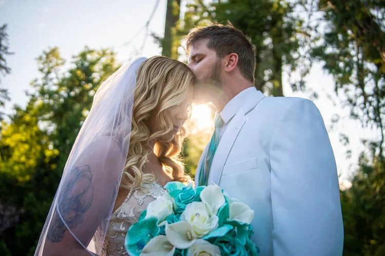 A bride and groom share a moment during their wedding, with the groom kissing the bride's forehead as she holds a bouquet of white and blue flowers, outdoors with green trees in the background.