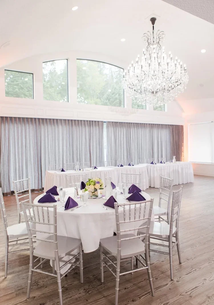 A banquet hall decorated for a formal event with white tablecloths, purple napkins, floral centerpieces, crystal chandelier, large windows, and curtains.