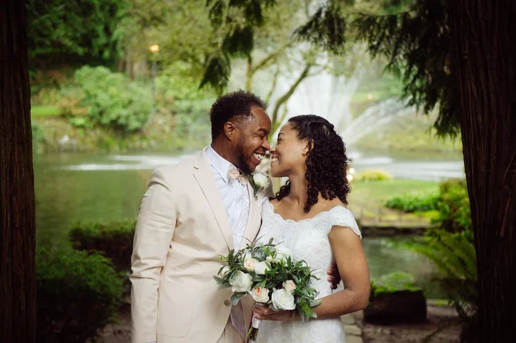 A newlywed couple smiling and touching foreheads in a lush, green outdoor setting near a waterfall and pond, with trees and foliage in the background.