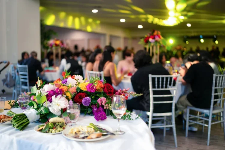 A banquet table decorated with a colorful floral centerpiece, set with plates, glasses, and food, in a large, well-lit event hall filled with seated guests.