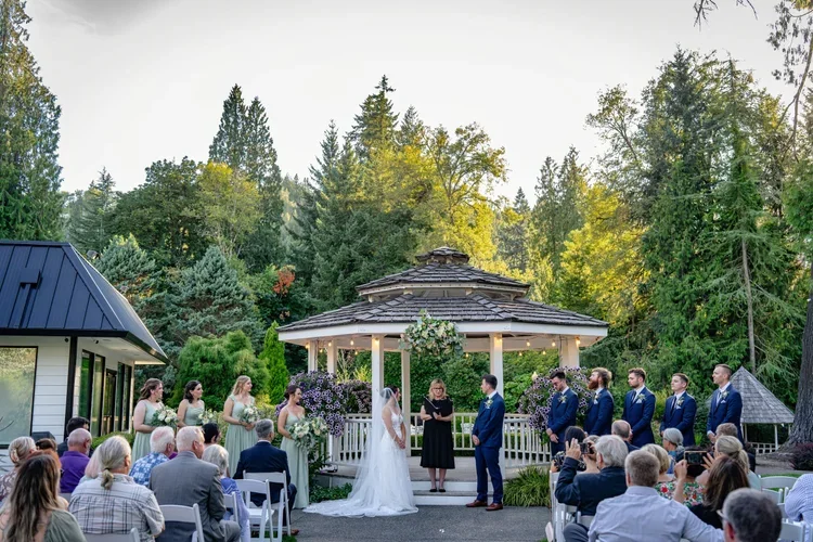 A wedding ceremony taking place outdoors in a garden setting with a gazebo, surrounded by trees. The bride and groom are standing together, facing an officiant, with the wedding party and guests seated on white chairs. The scene is during the daytime