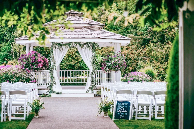 A white wedding gazebo decorated with white curtains and flowers, located outdoors among lush greenery with pink and purple flowers surrounding it, set for a wedding ceremony with rows of white chairs on either side of the aisle.
