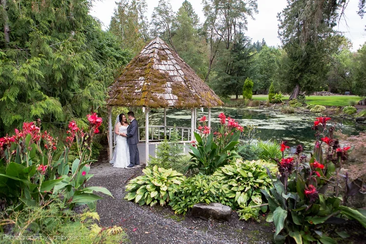 A wedding couple standing under a small rustic gazebo by a pond, surrounded by lush green plants and colorful flowers.