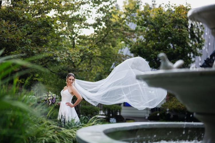Bride in a white wedding dress with flowing veil, standing in a lush garden with trees and greenery, holding a bouquet, with a fountain in the foreground.