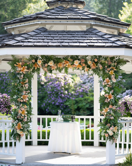 Wedding gazebo decorated with peach and white flowers and green foliage, with a small round table set with a decanter and glasses underneath.