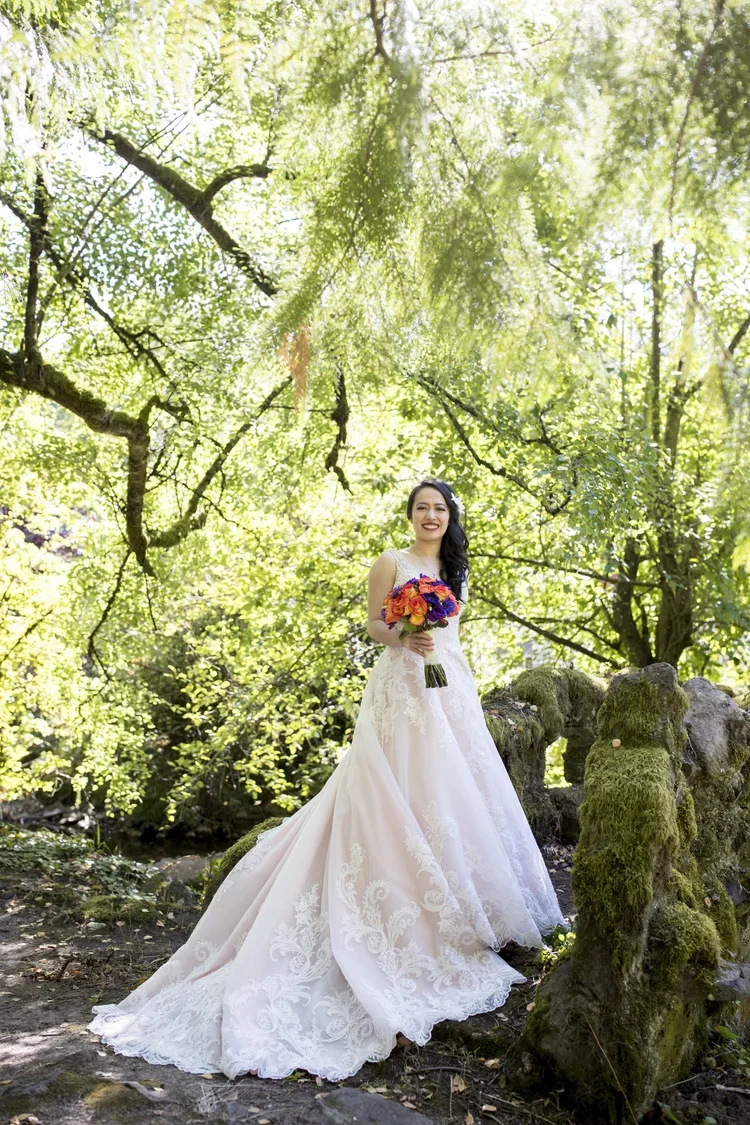 A woman in a white wedding dress holding a colorful bouquet, standing outdoors under green trees on a bright, sunny day.