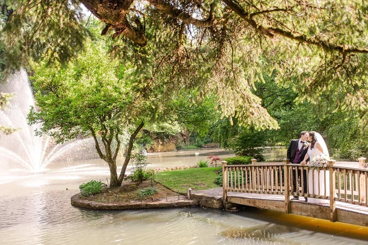 A newlywed couple sharing a kiss on a garden bridge over a pond, surrounded by lush trees and a fountain in the background.