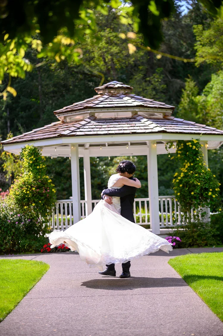 A bride and groom dancing outdoors under a gazebo, surrounded by green trees and colorful flowers.