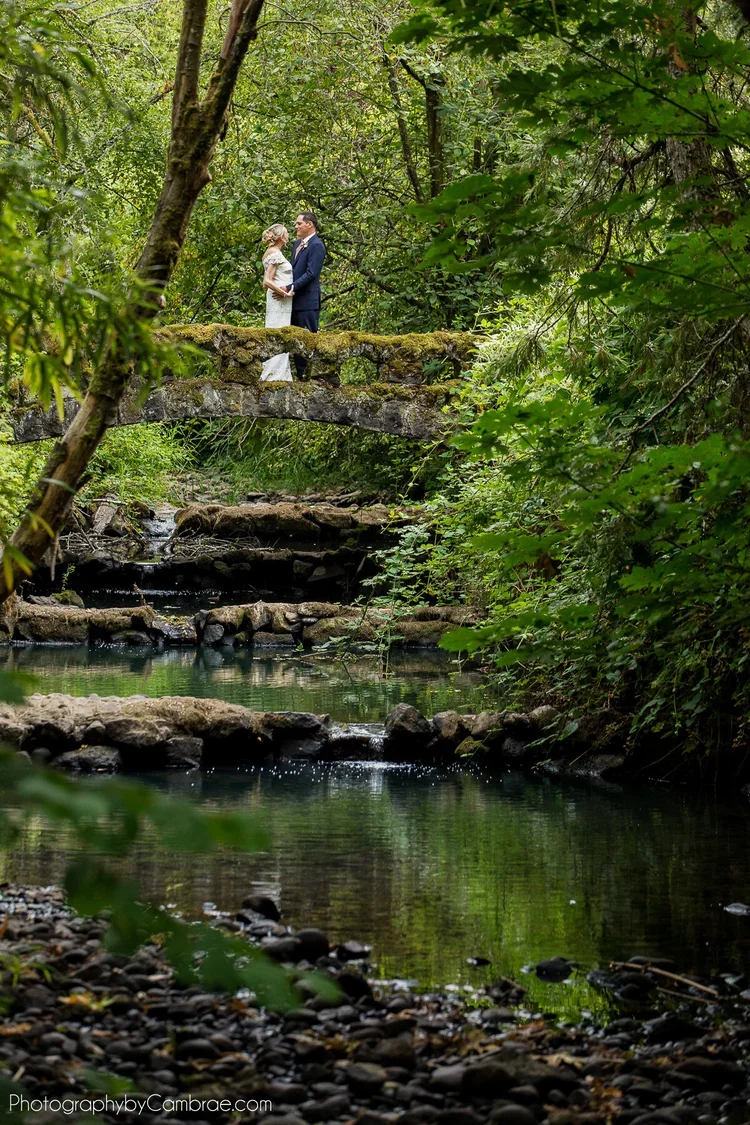 A couple dressed in wedding attire standing on a small stone bridge over a peaceful creek surrounded by lush green trees.