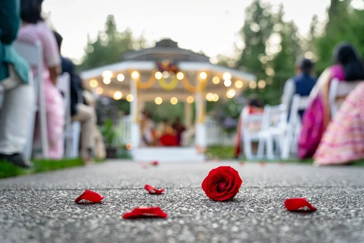 Red rose petals scattered on a gravel path at an outdoor wedding ceremony, with chairs and a decorated gazebo blurred in the background.