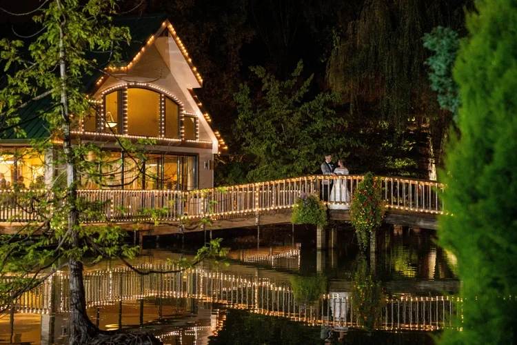 A night scene of a house with string lights on the roof and balcony, surrounded by trees, with their reflection visible on the water below where a couple dressed in formal attire is standing on the balcony.
