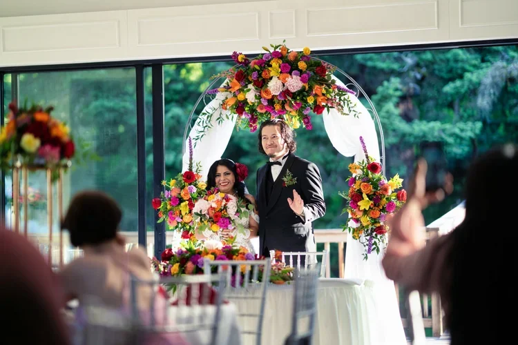 A bride and groom exchanging vows under a floral arch at an indoor wedding reception.