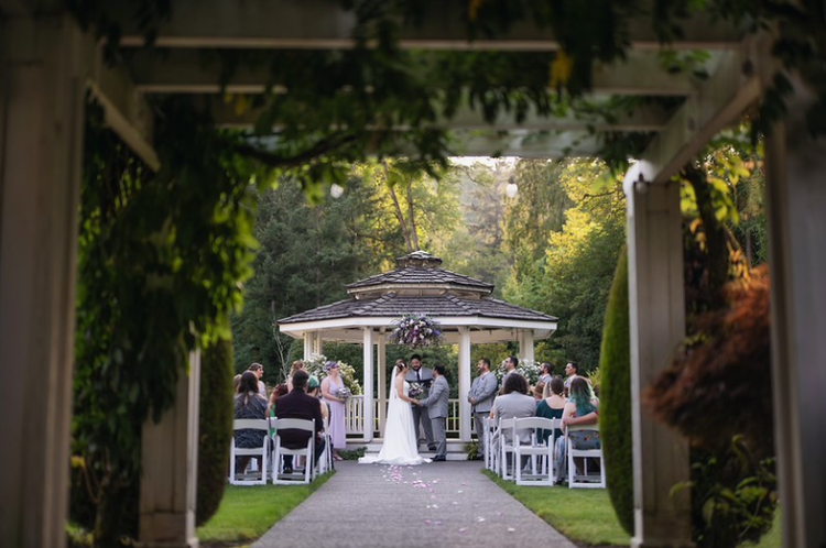 A wedding ceremony taking place outdoors under a pavilion, with the bride and groom standing in front of an officiant, surrounded by seated guests, in a garden setting.