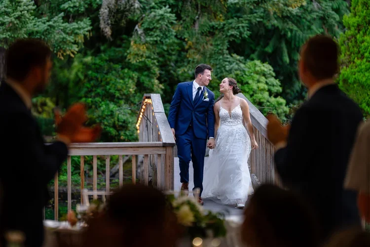 A newlywed couple holds hands and smiles at each other on a wooden bridge, with two people clapping in the foreground and lush green trees in the background.
