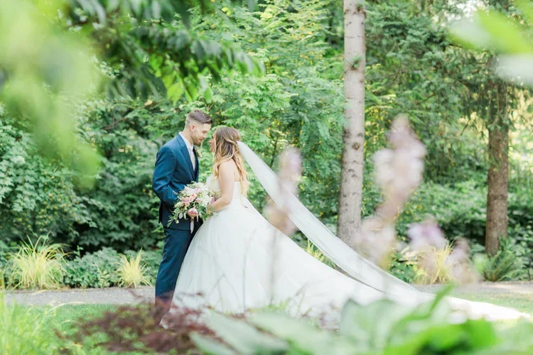 A bride and groom standing close together outdoors in a green, wooded area, sharing a tender moment on their wedding day.
