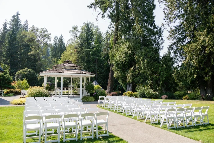 Outdoor wedding ceremony setup with white chairs arranged in rows, a white gazebo, lush green trees, and a clear sky in the background.