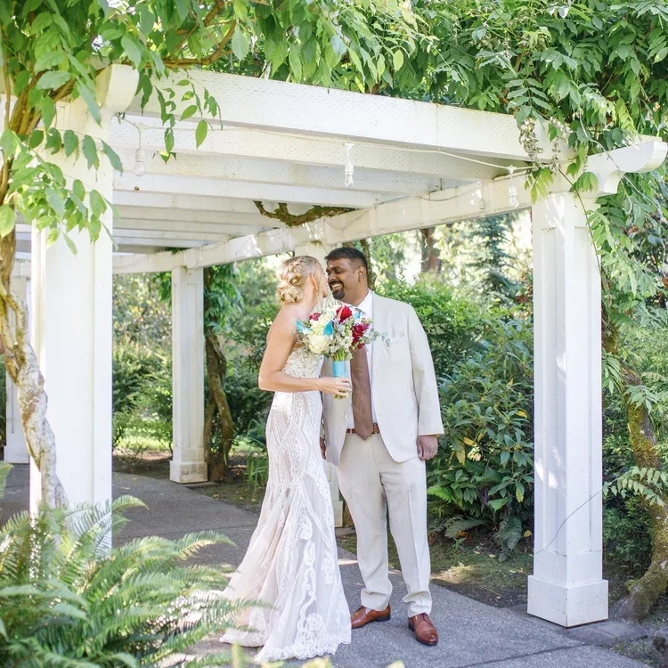 A newlywed couple standing under a white garden arbor with greenery around, the bride is wearing a white lace wedding dress and holding a bouquet, while the groom is in a light-colored suit and brown shoes, sharing a joyful moment.