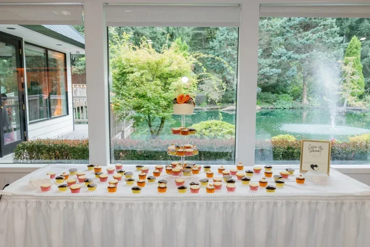 A table with a white tablecloth displaying assorted cupcakes with colorful frosting and toppings, set in front of large windows overlooking lush green trees and a pond outside.