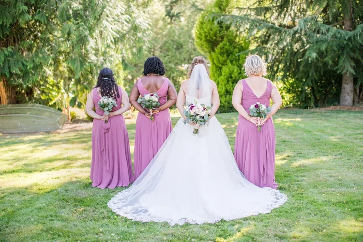 A bride and four bridesmaids standing outdoors in a lush, green garden, facing away from the camera, holding bouquets, with the bride wearing a white wedding gown and veil and the bridesmaids in matching purple dresses.
