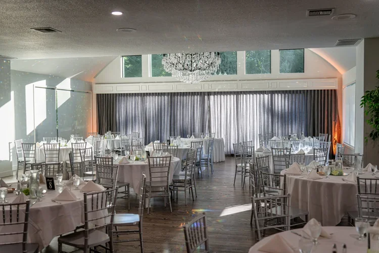An indoor banquet hall with round tables covered in white tablecloths, set with glassware and silverware, and surrounded by silver chairs. A large crystal chandelier hangs from the ceiling, and tall windows with gray curtains line the back wall. Sunl