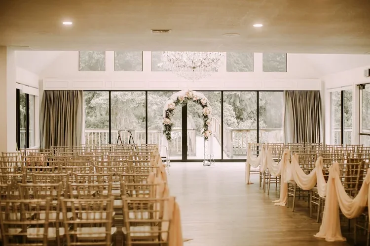 An indoor wedding venue with rows of chairs, a floral arch, and large windows overlooking trees.