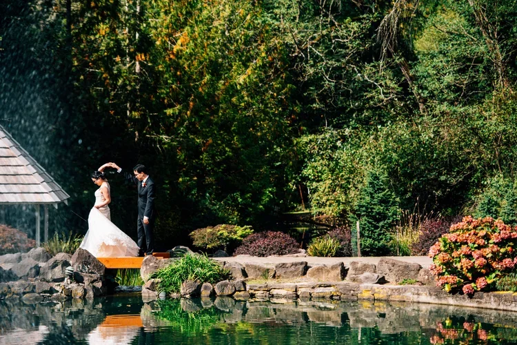 A bride and groom standing on rocks by a pond, surrounded by lush greenery and trees, during a wedding ceremony in an outdoor garden.