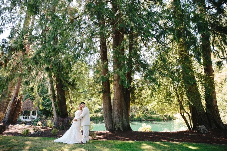 A bride and groom standing together in a park with tall trees, a pond, and greenery in the background, during a sunny day.