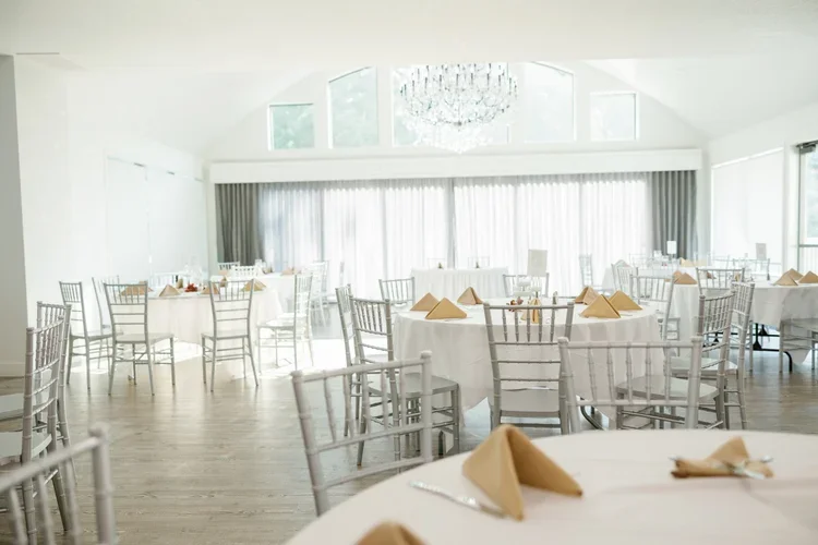 Elegant banquet hall with round tables covered in white tablecloths, beige napkins folded on each plate, silver chairs arranged around the tables, large windows with curtains, a crystal chandelier hanging from the ceiling, and a spacious bright atmos