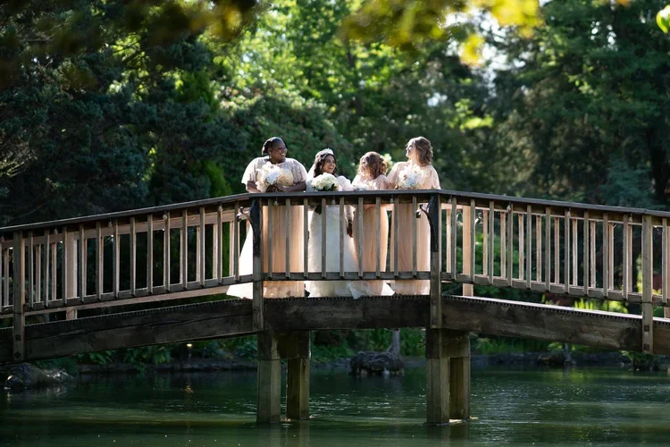 A group of five women dressed in wedding and bridesmaid gowns standing on a wooden bridge over a body of water, holding bouquets of flowers, surrounded by lush green trees and foliage.