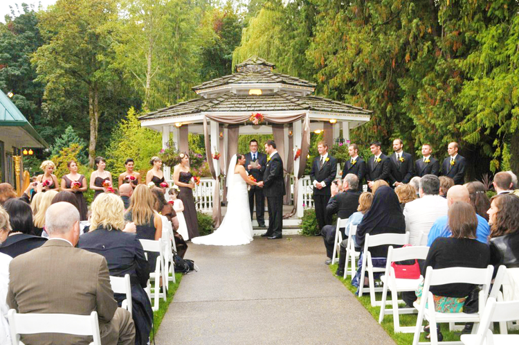 A wedding ceremony taking place outdoors under a gazebo, with a bride and groom exchanging vows, surrounded by their wedding party and seated guests, in a lush green setting.
