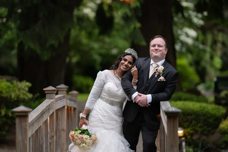 Happy bride and groom standing on a wooden bridge outdoors, smiling, with greenery and trees in the background.