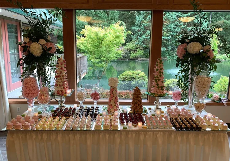 Dessert table with pink and white sweets, cakes, and candies, decorated with large flower arrangements, in front of large windows overlooking a lush green garden with trees and water.