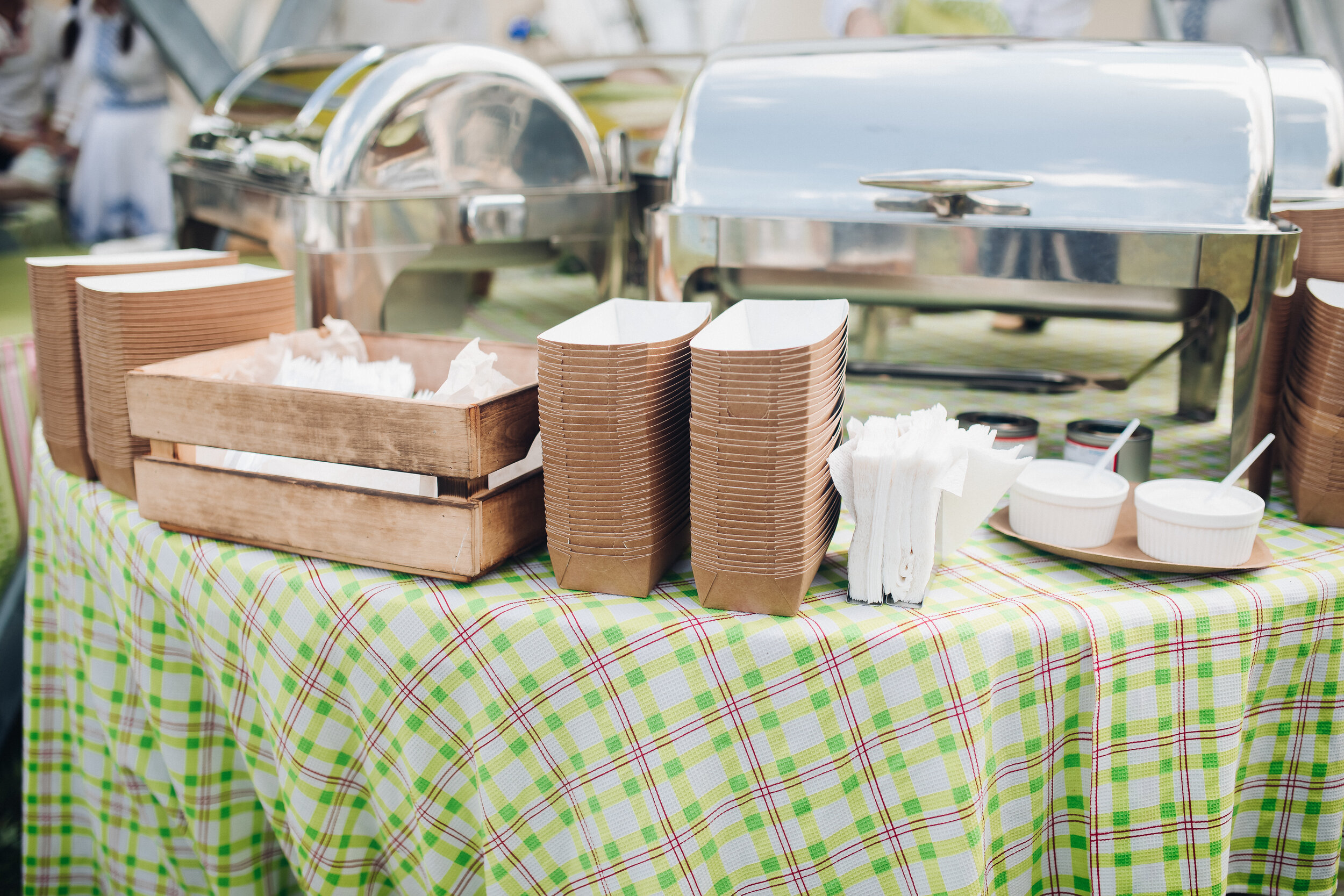Stock photo of paper recyclable containers in stacks on the table over patterned green and white tablecloth. Box of napkins and utensils. Outdoor food festival.
